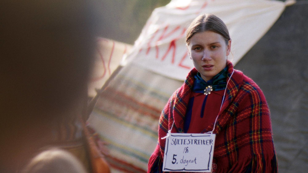 Ester (Ella Marie Hætta Isaksen) sit ved ein lavvo ved Stortinget. Ho er kledd i samisk kofte og sjal. Rundt halsen heng ein plakat som det står "Sultestreiker på 5. døgnet" på.